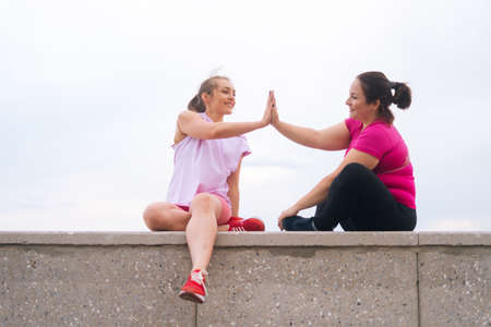Front view of cheerful slim fitness female trainer and motivated overweight young woman high-fiving each other after exercising outdoors. Instructor help fat woman lose weight outside.の写真素材