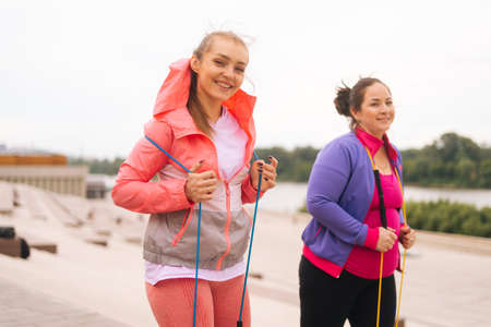 Medium shot of smiling overweight young woman doing exercises using fitness tape for weight loss with personal trainer outdoor in summer day. Fat female with coach stretching before running.の写真素材