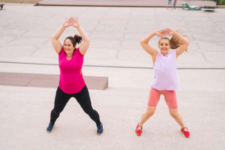 Front view of personal fitness female trainer giving training to positive overweight young woman performing jumping together, outdoor in summer day. Instructor help fat woman lose weight outside.の写真素材