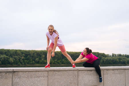 Wide shot of cheerful slim fitness female trainer and motivated overweight young woman fooling after exercising outdoors. Instructor help fat woman lose weight outside.の写真素材
