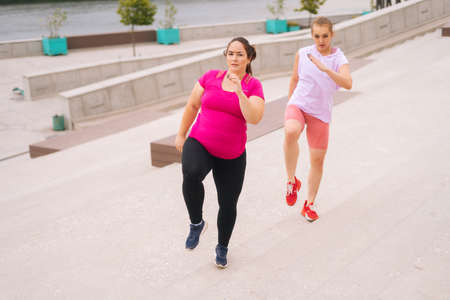 Personal fitness female trainer helping fat woman lose weight outside running on city stairs in summer morning. Instructor giving training to overweight young woman in cloudy summer day.の写真素材