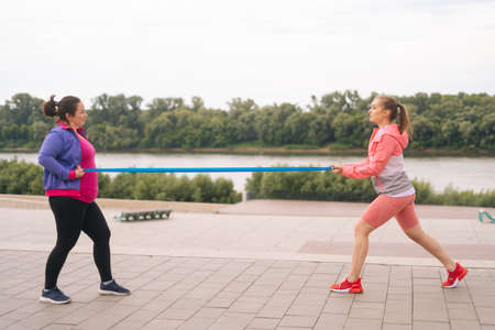 Wide shot of fat young woman doing squats exercises using resistance band for weight loss with personal trainer outdoor in cloudy summer evening. Instructor help overweight woman lose weight.の写真素材