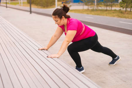 High-angle view of motivated fat young woman training doing push ups using street bench in city park at summer morning. Fitness female doing bench push ups for strength workout outdoors.の写真素材