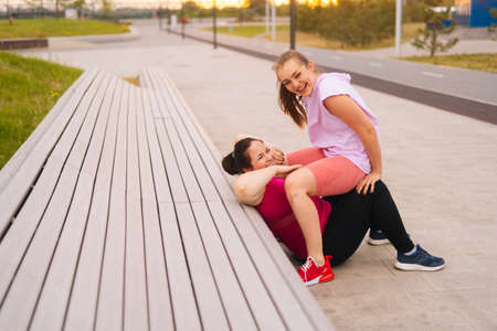 Cheerful fat young woman training with cheerful fitness instructor using bench at outdoors gym in city park. Personal training from athletic girl for happy fat young woman with big abdomen outsideの写真素材