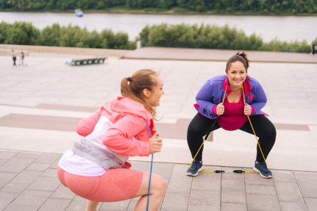 Wide shot of overweight young woman doing squats exercises using fitness tape for weight loss with personal trainer outdoor in summer day. Fat female with coach stretching before running.の写真素材