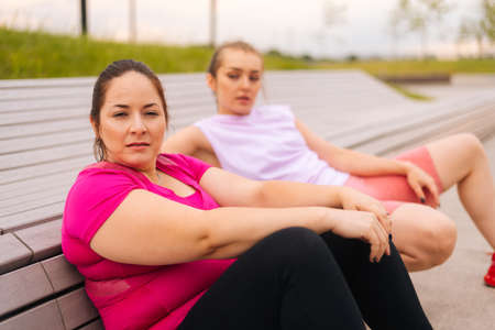 Close-up of tired obese young woman resting by bench after intense exercise with personal trainer outdoor in city park. Training from athletic lady for fat female with big abdomen wearing sportswear.の写真素材