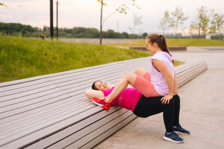 Positive obese young woman training with cheerful fitness instructor using bench at outdoors gym in city park. Personal training from athletic girl for happy fat young woman with big abdomen outside.の写真素材