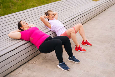 Side view of obese young woman exercising abdominal crunches using bench with personal trainer outdoor in summer morning. Training from athletic lady for fat female with big abdomen wearing sportswearの写真素材
