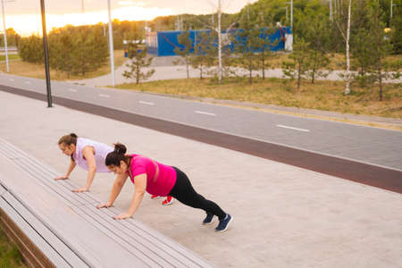Wide shot of personal training from athletic lady for obese female with big abdomen outdoor in cloudy summer evening. Fat caucasian woman performing push up exercise with female trainer using bench.の写真素材