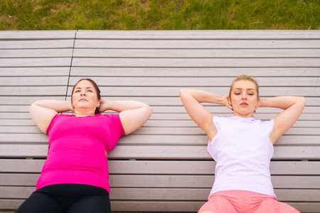 Top view of obese young woman exercising abdominal crunches using bench with personal trainer outdoor in summer morning. Training from athletic lady for fat female with big abdomen wearing sportswear.の写真素材