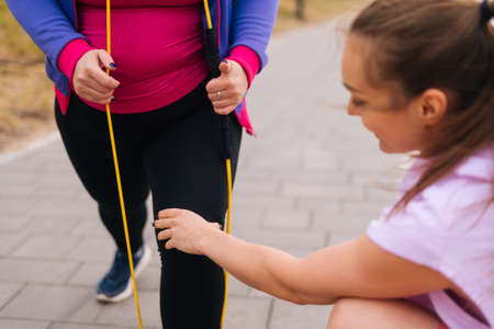 Close-up cropped shot obese overweight young woman doing exercises using fitness tape for weight loss with personal trainer outdoor in summer day. Fat female with coach stretching before running.の写真素材
