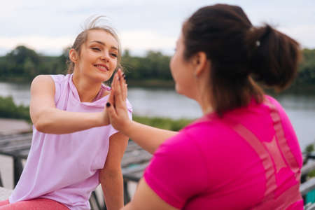 Close-up back view of positive fitness female trainer and motivated fat young woman greeting each other with high-five other after exercising outdoors. Instructor help obese woman lose weight.の写真素材