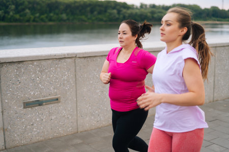 Side view of personal fitness female trainer helping motivated fat woman lose weight outside running along city waterfront in morning. Instructor giving training to overweight young woman outside.の写真素材