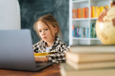 Portrait of serious pensive focused primary child school girl doing homework sitting at table with...