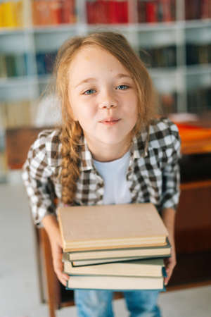 Vertical portrait of playful elementary child school girl holding stack of books in library at...