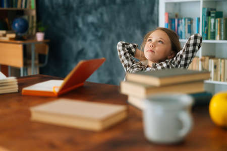 Side view of relaxed happy pupil school girl kid sitting with paper book and notebook at desk,...