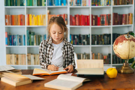 Front view of attractive pupil school girl kid doing homework writing in notebook sitting at table with paper book. Portrait elementary child schoolgirl studying at home, selective focus.の写真素材