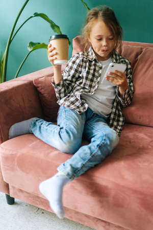 Vertical shot portrait of adorable cute little child girl using smartphone looking at screen, holding coffee cup in hands, sitting on couch at home alone, playing mobile device game.の写真素材