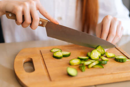 Close-up cropped shot of unrecognizable woman cutting fresh cucumber cooking food salad sitting at wodden table in light kitchen room. Side view of female cooking vegetarian dieting salad at homeの写真素材