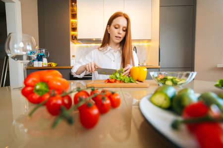 Front low-angle view of young redhead woman cutting fresh lettuce preparing food salad sitting at table in modern kitchen room. Pretty female cooking vegetarian dieting salad full of vitamins.の写真素材
