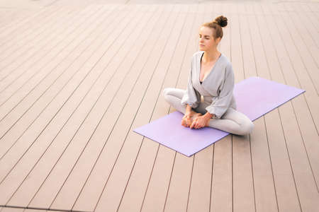 Wide shot of calm young woman sitting on yoga mat with closed eyes and meditating in butterfly pose,...