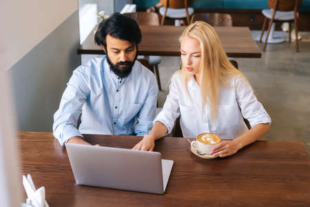 High-angle view of two serious male and female business colleagues sitting at table and talking in cafe looking at laptop screen. Focused people talking during business meeting in coffee shop.の写真素材