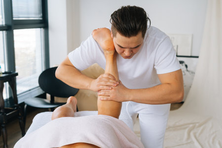 Front view of professional male masseur with strong hands massaging calf muscles on lower leg of muscular athlete man after physical sports workout, lying on stomach at massage table.の写真素材