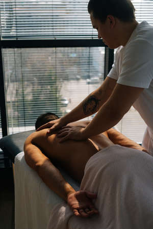 Vertical back view of professional male masseur with strong tattooed hands massaging back and shoulders of muscular sports man lying on stomach, at massage table on background of window.の写真素材