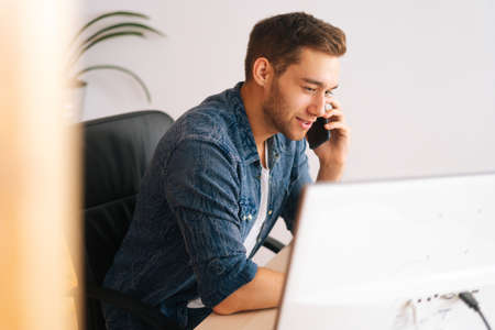 Side view of focused young business man talking on mobile phone sitting at desk with monitor...