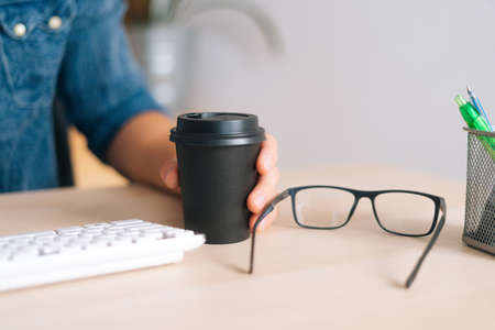 Close-up of male hands holding cup with coffee sitting at wooden working desk with stylish eyeglasses, wireless white keyboard and different chancellery laying around, selective focus, coffee break.の写真素材