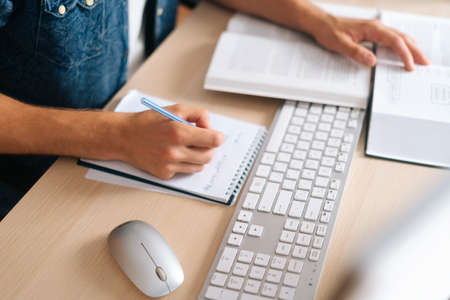 Close-up top view of unrecognizable male student studying online from home using computer, writing...