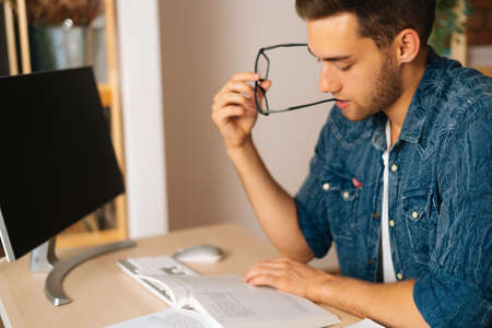 Close-up side view of focused thinking young man reading paper book holding end of glasses near...