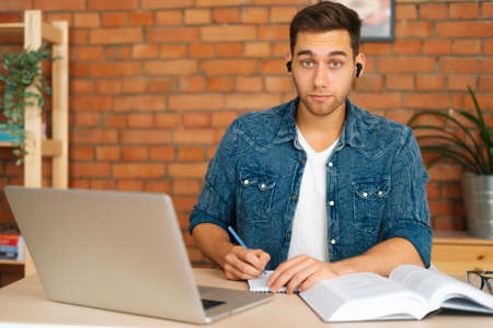 Front view of serious male student studying online from home using laptop, writing notes, watching video class lesson, listening audio course, studying with teacher distant, lookign at camera.の写真素材