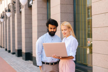 Medium shot portrait of two skilled businesspeople talking about project holding laptop standing in...