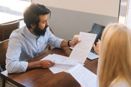 High-angle view of two diverse colleagues discuss financial data in report using paper documents and...