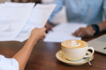 Close-up of unrecognizable businessman and businesswoman discussing and checking paper documents, sitting at table in cafe with cup of hot latte coffee with beautiful pattern, selective focus.の写真素材