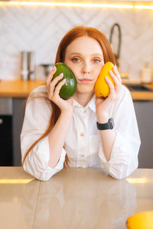 Vertical portrait of serious attractive redhead young woman holding in hands mango and avocado...