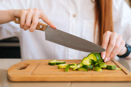 Close-up front view of unrecognizable woman cutting fresh cucumber cooking food salad sitting at wodden table in light kitchen room. Front view of female cooking vegetarian dieting salad at home.の写真素材