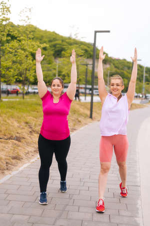 Full length vertical portrait of fitness female trainer have personal training with overweight young woman outdoor in morning. Instructor help fat woman lose weight outside practising yoga asana.の写真素材