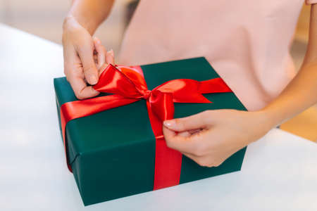 Close-up top view of unrecognizable female hands wrapping Christmas gift box tying red ribbon and decorating beautiful bow, closeup. Xmas or New year gift in stylish wrapping paper with red ribbon.の写真素材