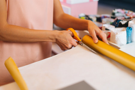 Close-up top view of unrecognizable young woman cutting with scissors wrapping golden paper to package festive cardboard box with Christmas present. Concept of new year and xmas, preparing presents.の写真素材