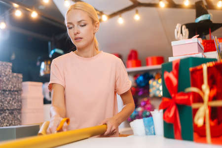 Low-angle view of young woman cutting with scissors wrapping golden paper to package festive cardboard box with Christmas gifts, at counter of holiday store, blurred background of festive packages.の写真素材