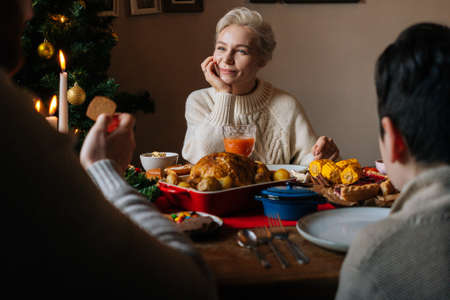Close-up view from back to happy young woman in love smiling while listening to husband and son at festive Christmas dinner table, during holiday family party, eating and chatting.の写真素材