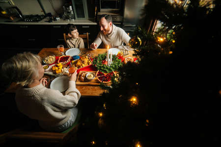 High-angle view of happy young family enjoying at festive Christmas table during holiday family party. Cheerful mother, father and little son having dinner xmas party, eating and chatting at home.の写真素材