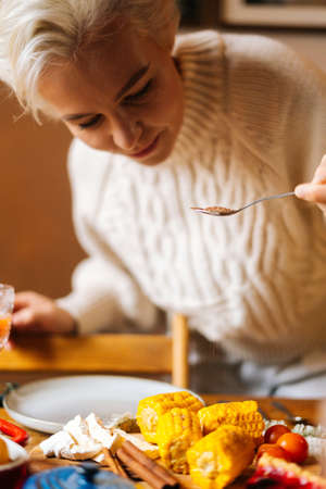 Close-up vertical shot of young woman preparing Christmas dinner table at home for family party, sprinkles boiled corn with spices. Pretty female serving tableware waiting for friends.の写真素材