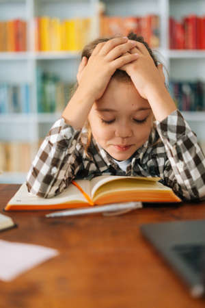 Close-up vertical shot of exhausted primary child school girl tired from studying holding head head with hands while reading book sitting at desk with workbook. Child schoolgirl doing homework at homeの写真素材