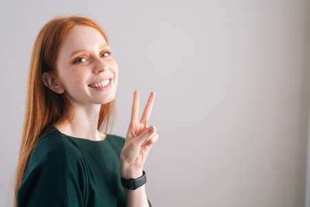 Portrait of smiling redhead young woman model showing victory sign with fingers looking at camera on white background. Medium shot of red-haired lady with natural beauty posing in professional studio.の写真素材