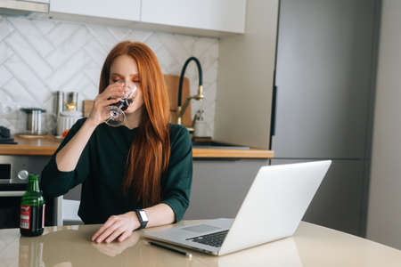Attractive redhead young woman drinking alcohol beverage in glass sitting at desk with laptop in kitchen with modern light interior. Pretty happy female freelancer drinking wine alone at home.の写真素材