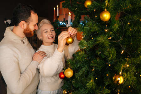 Happy young couple in love decorating Christmas tree with colorful shiny balls near fireplace in cozy dark living room. Cheerful man and woman preparing home for New Year holidays celebrationの写真素材