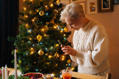 Side view of pretty blonde young woman taking photo or making video of beautiful Christmas dinner table on smartphone, on blurred background of decorating xmas tree and celebration bokeh lights.の写真素材